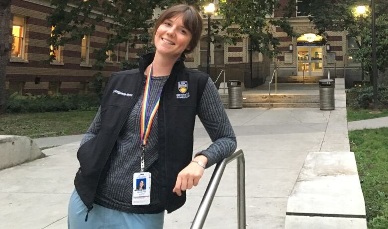 Dr. Marguerite Hanes stands in front of a hospital in scrubs and a black vest. She's leaning on a handrail and looking at the camera.