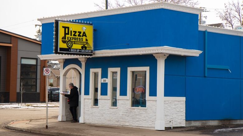 A person walks out of a blue building carrying pizza boxes.