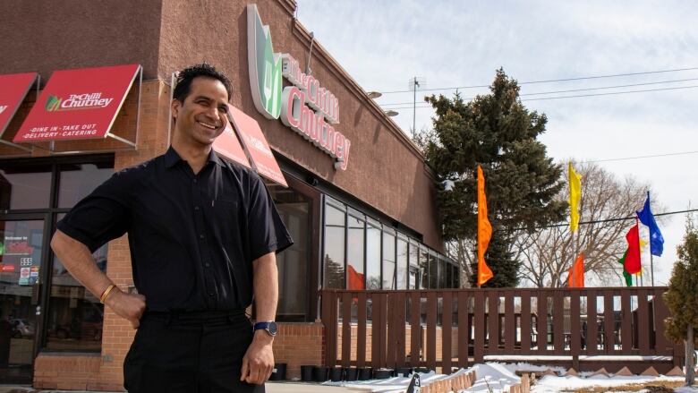 A man dressed in black stands smiling outside an Indian restuarant.