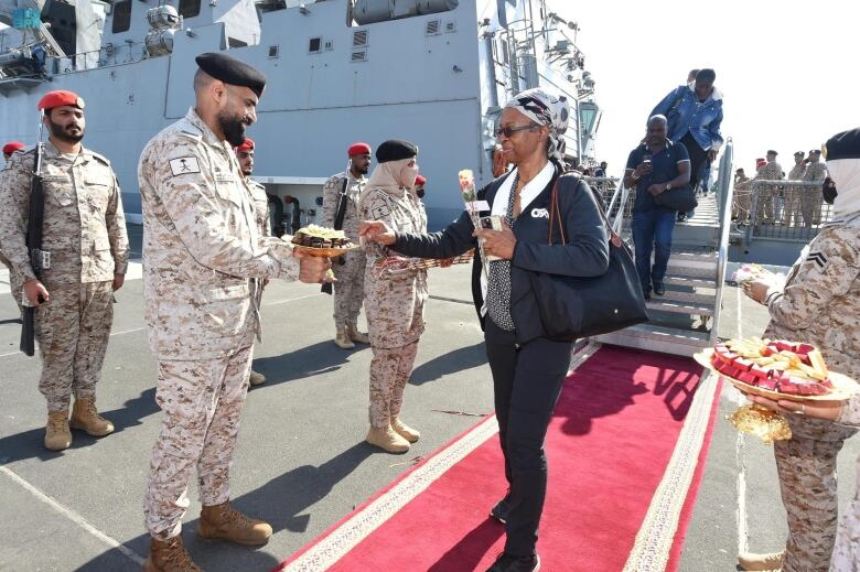 A woman holding a flower stands on a red carpet, lined by men in brown camouflage uniforms, in front of a grey naval vessel. 