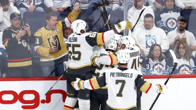 A group of male ice hockey players wrap their arms around each other while celebrating against the boards as fans of both teams look on from behind the glass.