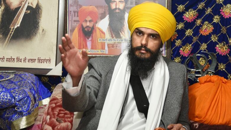 A man in a yellow turban, a long beard and a neutral expression gestures with his right hand.