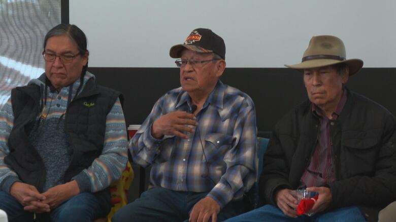 Three men are seated and appear to be talking to a crowd.