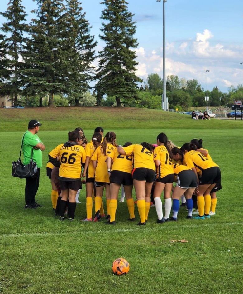 Girls wearing bright yellow t-shirts, black shorts, and knee-high yellow socks, stand in a huddle turned away from the camera. They are on a green soccer pitch and there is an orange soccer ball on the pitch. 