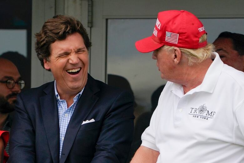 Two men are seen in conversation, one is wearing a baseball cap.