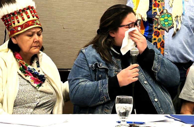 A woman in a jean jacket wipes away tears with a tissue while another woman comforts her. 