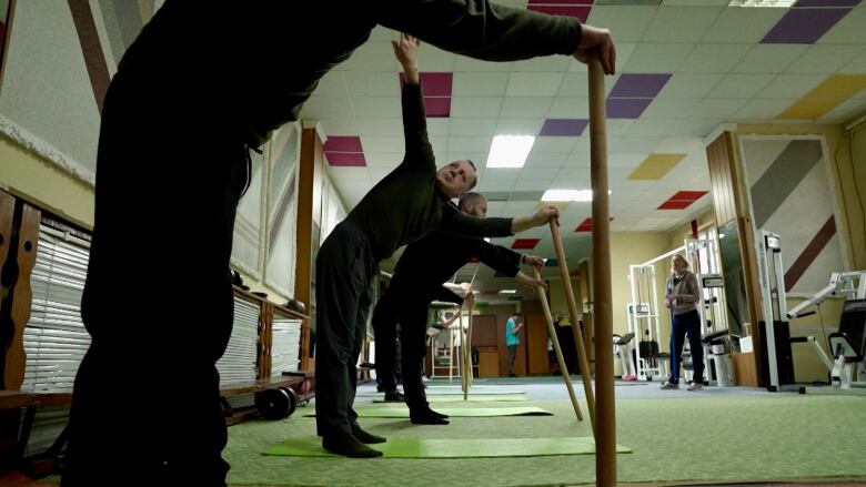 Men perform stretching exercises in a gym while a woman watches. 