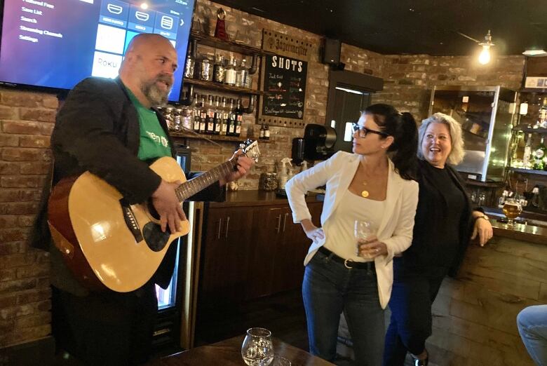 A man, left, plays guitar. Two women, one wearing white and the other in black, stand to the side and watch.