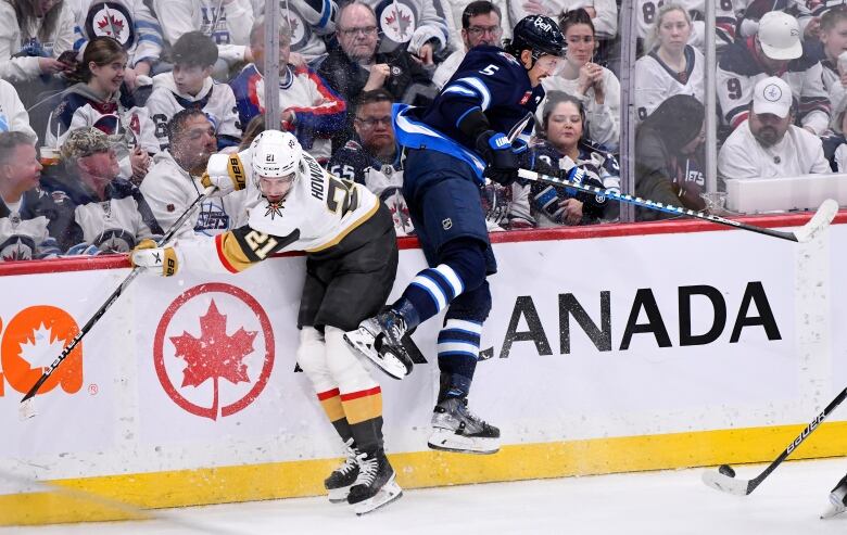 A Winnipeg Jet, in blue, jumps to avoid a hit along the boards from a Golden Knight, in white.