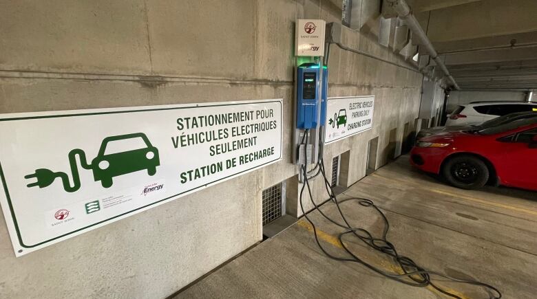 A sign with a car and plug symbol. The sign is next to a blue charging station on the wall. There are two cars parked one parking space away from the charger.