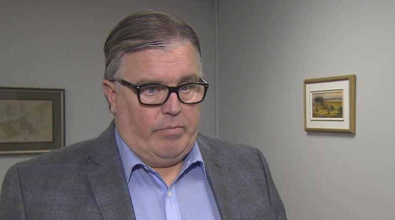 A grey haired man in glasses wearing a suit jacket looks off camera