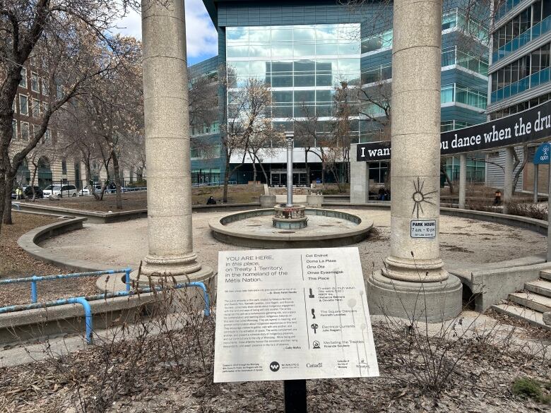 A park can be seen with tall concrete pillars, concrete steps and a central concrete fountain. The fountain is empty. 