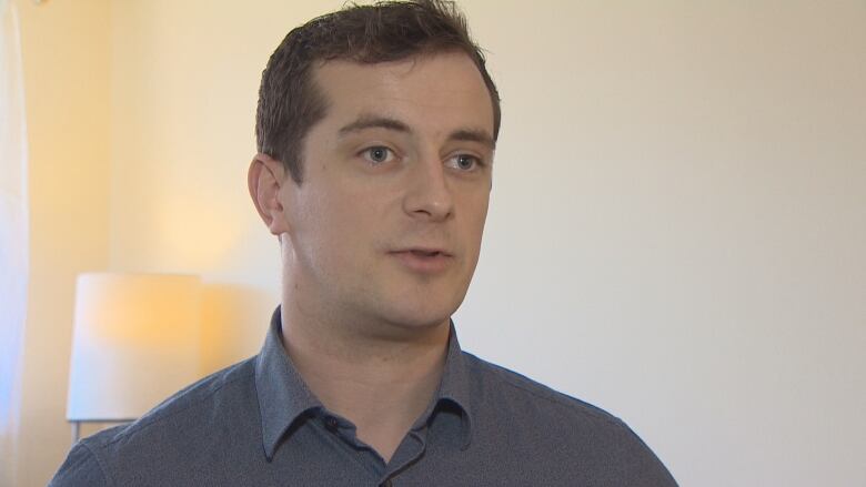 A man with dark hair is seen wearing a blue-grey button-up shirt. 