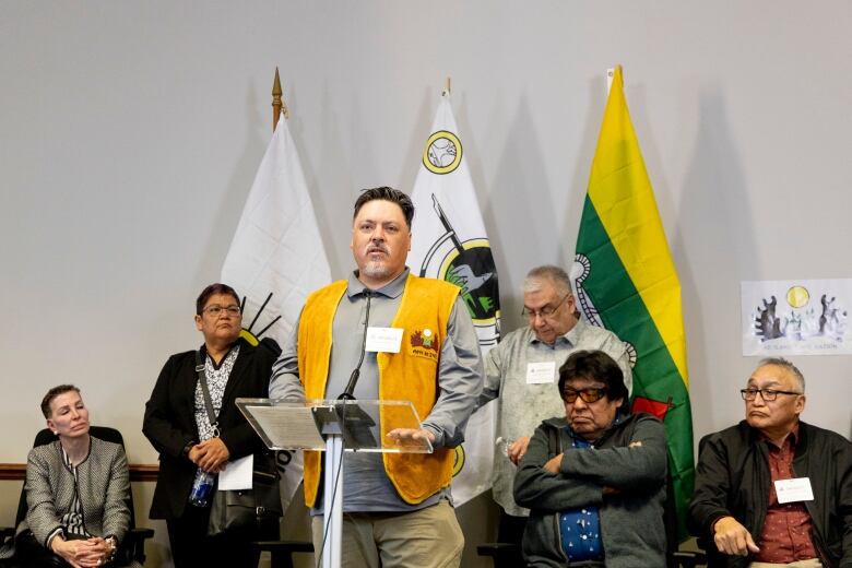 A man stands up and speaks at a microphone, while people sit behind him listening. 