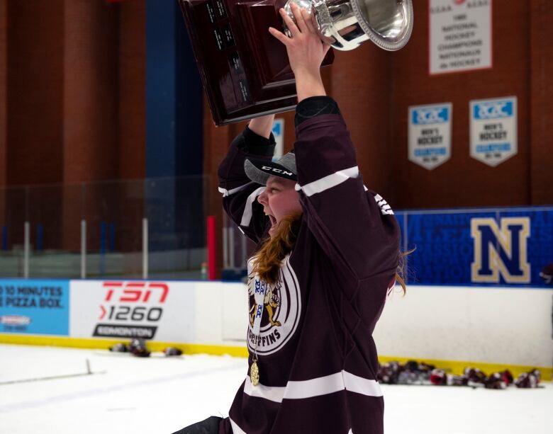 A woman raises a trophy over her head on the ice surface of a hockey arena.