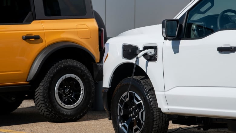 The front end of a white pickup truck is pictured with a charging cable plugged in above the driver's side front tire. 