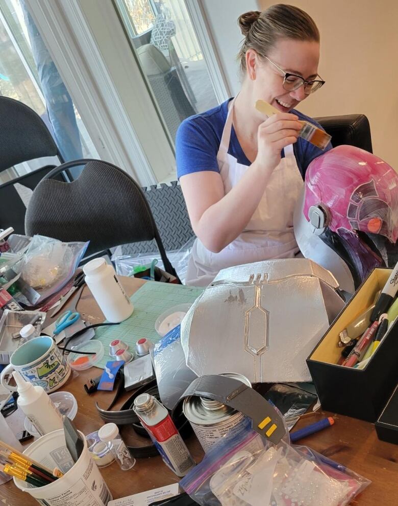 A woman sits at a table covered in craft supplies painting a helmet.