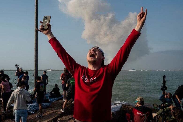 A man in a red shirt stands cheering, with his arms outstretched and his face looking to the sky, as a cloud of smoke from a space launch rises in the background.