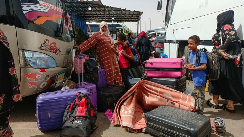 A family looks over a pile of luggage as they stand between two busses under a hot sun.  People and other vehicles are pictured in the background. 