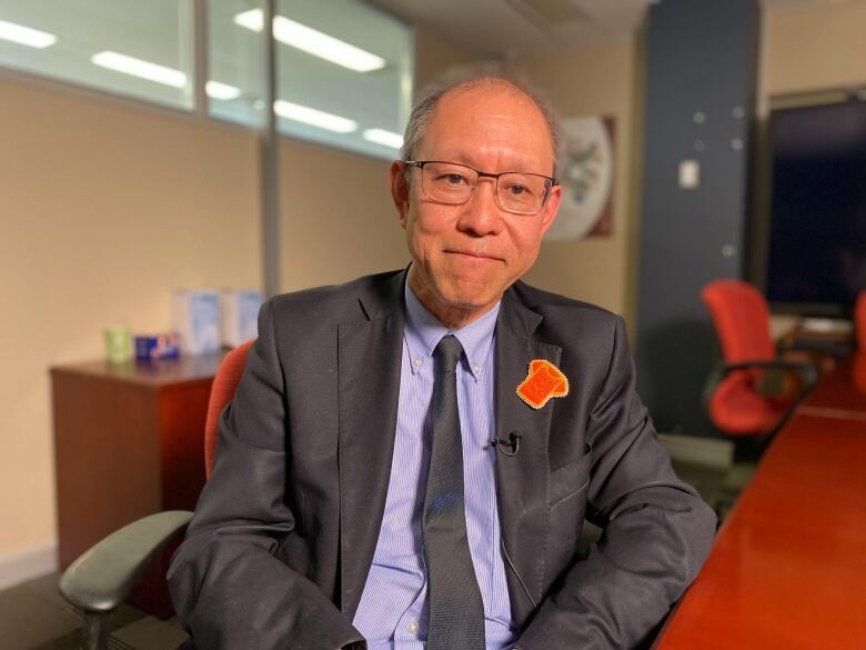 A man in a suit looks into the camera in an office boardroom. He wears an beaded orange shirt pin on his left lapel.