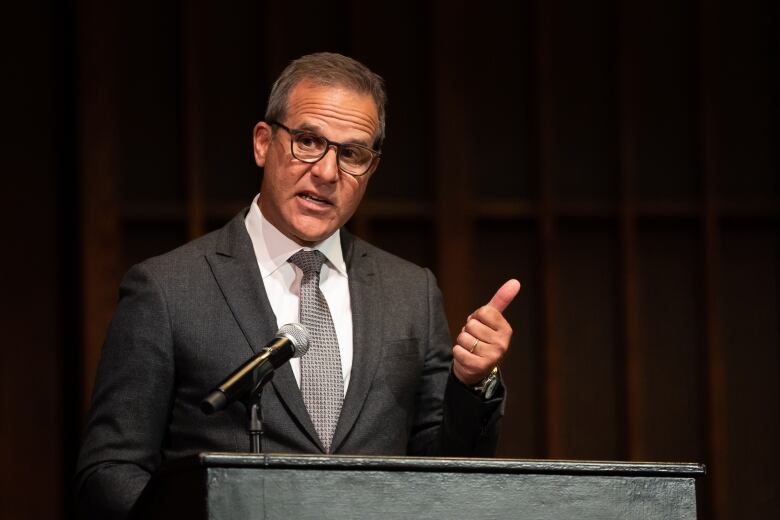 Jordan Bitove on stage delivering a lecture. He is at a podium, wearing a suit and gesturing with his hand with his thumb up. 