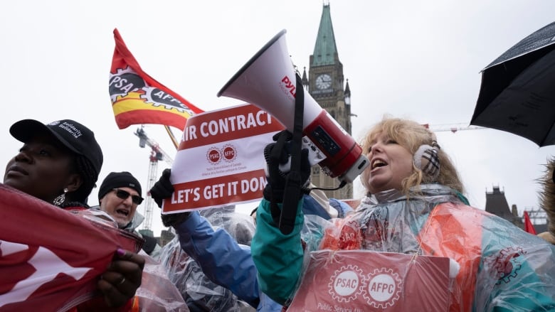 Striking government workers, including one with a megaphone, rally in front of a legislature on a drizzly spring day.