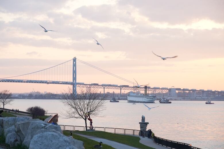 A photo of a steamship dating to 1907 being tugged through the river, with the Ambassador Bridge and sunset in the background. 