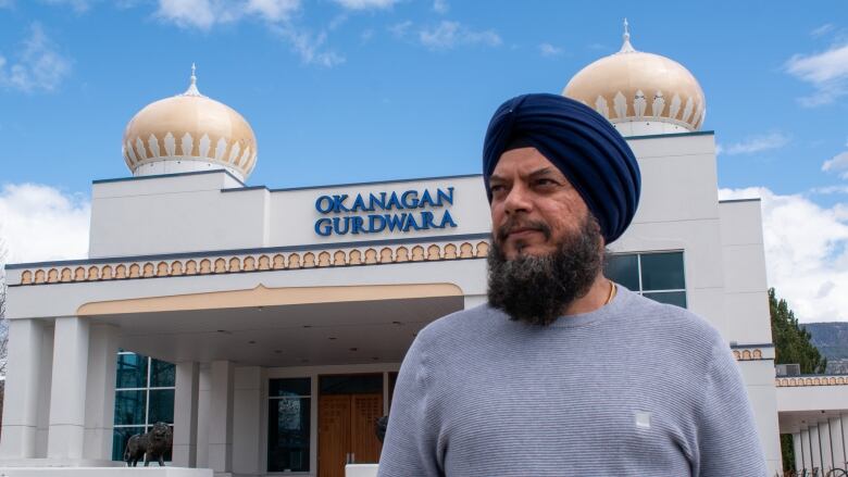 A man with blue turban and grey shirt stands in front of a building with logo 'Okanagan Gurdwara.'