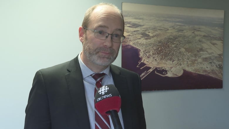 A balding man in a suit and tie stands in front of a microphone with a CBC gem on it