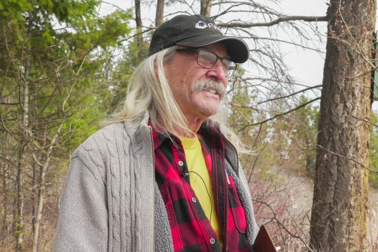 A man with long white hair and a mustache under a baseball cap wearing a beige sweater over a red lumberjack shirt and a yellow T-shirt, with a forested background behind him, smiles 