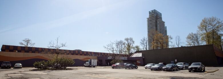 A large parking lot with a few cards and a long flat building with a high rise in the background. 