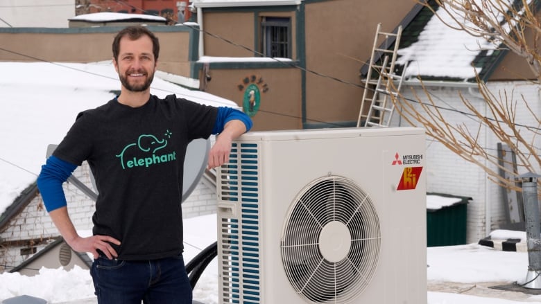 A man leans on the outdoor unit of a heat pump with snow in the background.