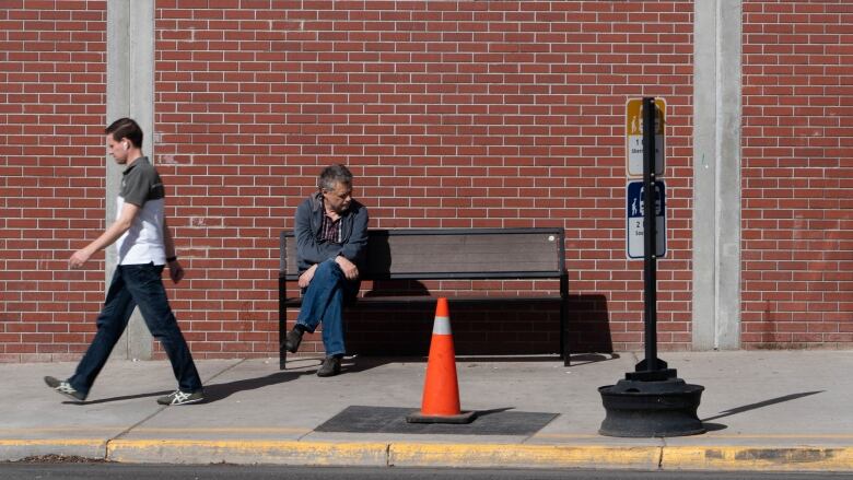 A man sits on a bench in front of a brick building next to bus signage as another walks past him