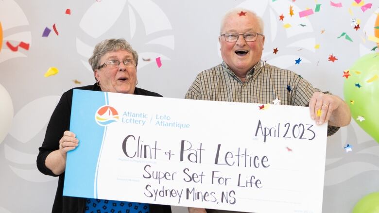 An older couple holds a large fake cheque with their names on it as balloons and confetti fall around them.