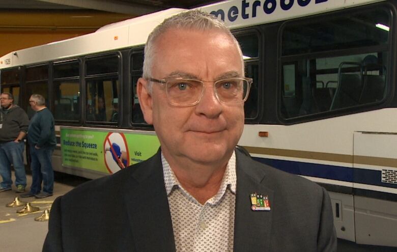 A man with glasses and wearing a black suit jacket and white shirt standing in front of a public bus. 