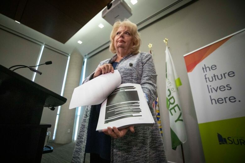 A woman with blonde shoulder-length hair standing at a podium holds open a report showing a page with blocks of blacked-out text.