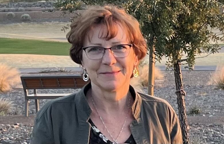 A woman smiles in an outdoor setting in front of a tree and a bench.