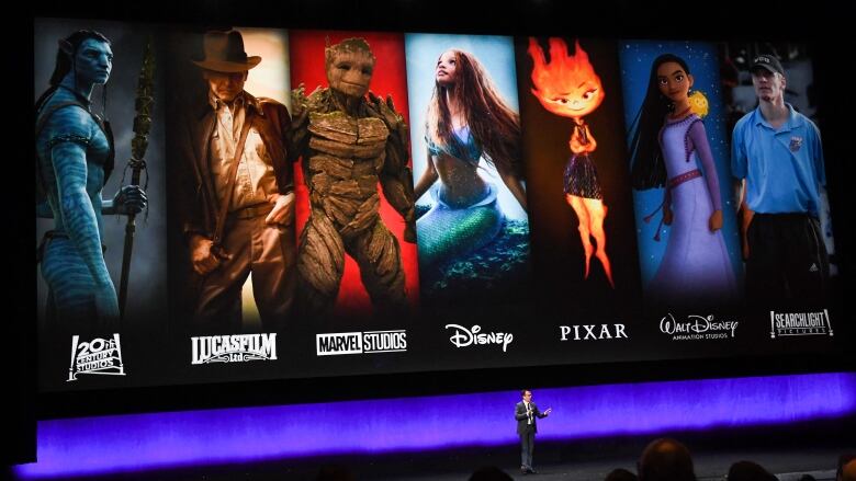 A man appears on a stage with a large screen behind him showing characters from various films.