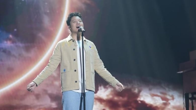 Teenage boy wearing a beige jacket sings into a mic with his arms stretched to the side. Behind him is a red, black and yellow screen. 