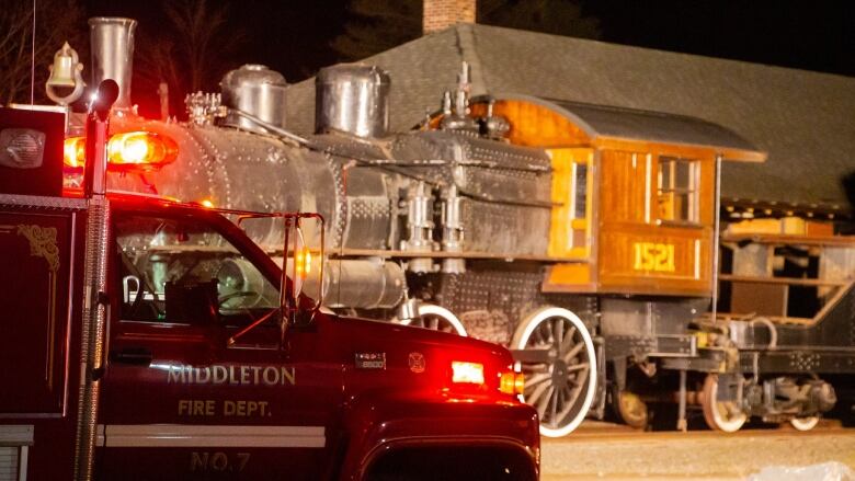 A fire truck is parked in front of an old train car and engine with its lights on.