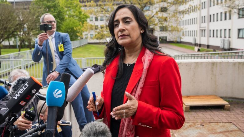 A woman in a long red coat gestures as she speaks at a phalanx of microphones.