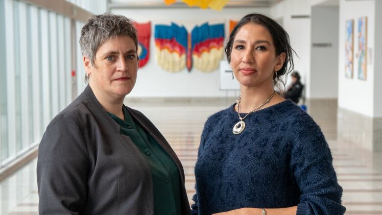 Two women stand in a hallway with serious facial expressions.