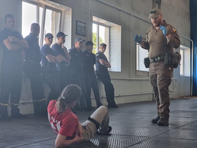 A man wearing a uniform holds his hands in air over someone on the ground, as people watch from the side, during a training exercise.