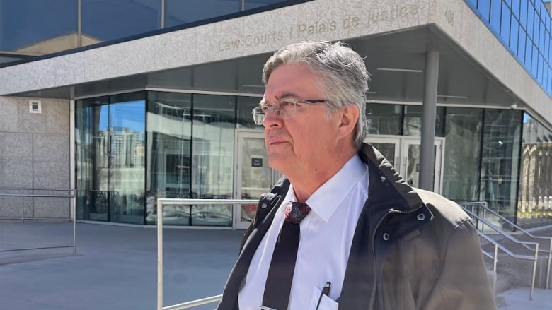 A man with grey hair and wearing glasses walks past a building as the sun shines. He has a light spring jacket, white collared shirt and black tie.