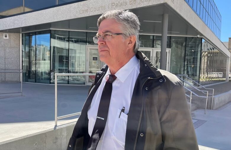 A man with grey hair and wearing glasses walks past a building as the sun shines. He has a light spring jacket, white collared shirt and black tie.