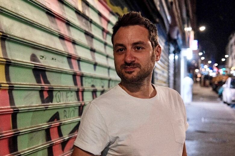 A man stands in an alley at night in front of a garage door covered in graffiti. 