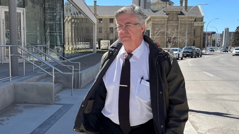 A man with grey hair and wearing glasses walks past a building as the sun shines. He has a light spring jacket, white collared shirt and black tie.