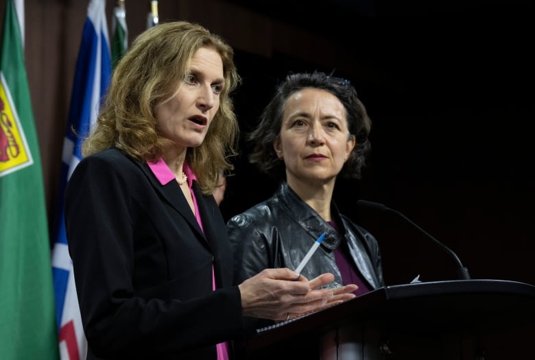 A women in a black suit stands at a podium and answers media questions as another woman in a leather jacket looks on.