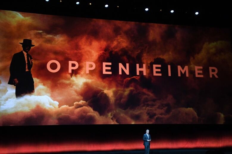 A man speaking in front of a large display of a movie poster. 