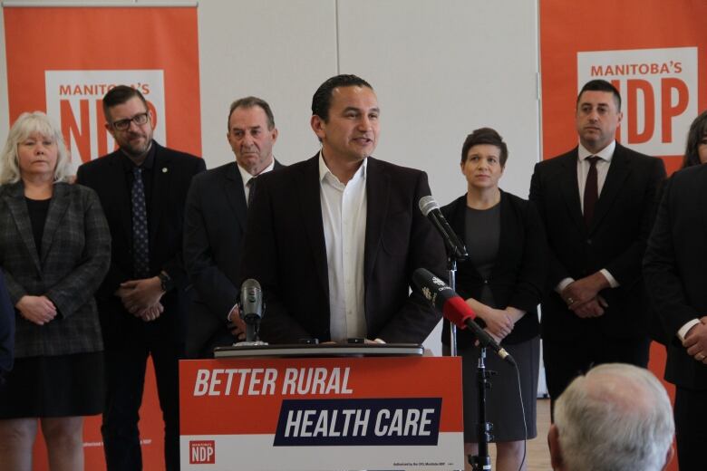 A man in a white collared shirt and dark blazer speaks to a crowd at an election campaign announcement.